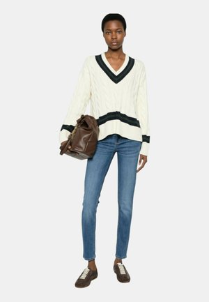 Donna che indossa un maglione bianco a trecce con strisce nere, jeans blu, sneaker marroni, che tiene una grande borsa in pelle marrone.