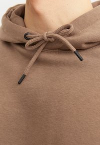 Il pullover hoodie marrone presenta un cappuccio con coulisse e texture intrecciata, punte nere sulla coulisse e una finitura in tessuto liscia.