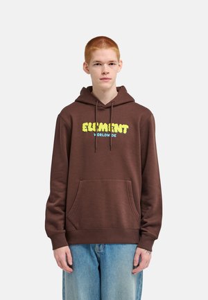 Giovane con capelli rossi e corti indossa una felpa marrone con la scritta “Element Worldwide” e jeans blu, in piedi davanti a uno sfondo chiaro e semplice.