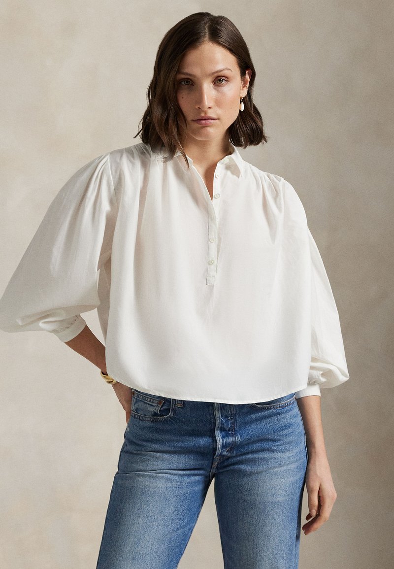 Hvit bluse med knapper, pufferm, lett stoff, avslappet passform og avrundet kant, kombinert med blå høyhælte jeans.