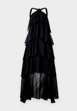 Robe maxi noire à dos nu avec volants superposés, texture légère et transparente, et une silhouette longue et fluide. Détail en nœud au niveau du cou.