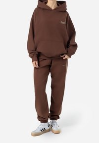 Sudadera marrón con un bolsillo frontal y logo, combinada con pantalones jogger a juego. Lleva zapatillas blancas con rayas negras y acentos beige.
