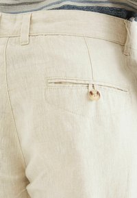 Lyse beige linnedbukser har en baglomme med en rund, lys brun knap og flade syninger. Diskret tekstur er synlig i stoffet.