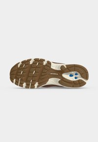 Καφέ και λευκή σόλα παπουτσιού New Balance με υφή και τρεις μικρές μπλε εμφανίσεις κοντά στη φτέρνα σε ανοιχτό γκρι φόντο.
