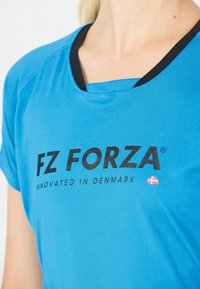 Närbild av en person som bär en blå FZ FORZA sporttröja med svarta detaljer och texten "Innovated in Denmark" samt en liten dansk flagga.