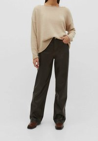 Beżowy, oversize sweter z zaokrąglonym dołem, noszony na ciemno-brązowych spodniacach o szerokim kroju. Materiał wydaje się miękki z teksturowanym splotem.