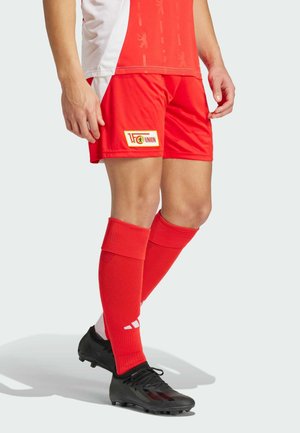 Rode voetbalkorte broeken met witte accenten, voorzien van een teamlogo patch. Zwarte voetbalschoenen en rode sokken maken de sportieve look compleet.