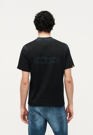 Άνδρας με σγουρά μαλλιά που φοράει μαύρο μπλουζάκι Replay Inc. και μπλε τζιν, εμφανίζεται από πίσω σε απλό φόντο.