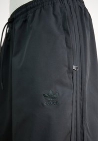 Schwarze Sportshorts mit einem elastischen Bund, seitlichen Reißverschlusstaschen und einem gestickten grünen Adidas-Logo auf der unteren linken Seite. Glatte Textur.