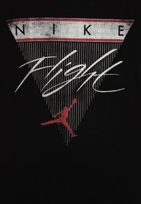 Černé tričko s grafickým designem, na kterém je v bílé a červené barvě napsáno "NIKE", skákající postava v červené a vertikální bílé čáry tvořící trojúhelník.