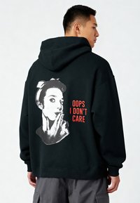 Sudadera con capucha negra con un gran gráfico blanco de una cara haciendo un gesto con la mano, y el texto "OOPS I DON’T CARE" en rojo en la parte trasera. Material de algodón.
