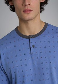 Camicia blu a maniche lunghe con colletto grigio, due bottoni e un motivo di piccoli pattini e fiori rossi. Realizzata in morbido cotone, dal design casual.