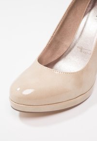 Chaussure à talons hauts pour femme en cuir verni beige avec bout arrondi et semelle intérieure métallique argentée rembourrée sur fond blanc.