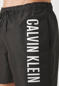 Crne kupaće gaće od laganog materijala s elastičnom traseom, s istaknutim bijelim logotipom "CALVIN KLEIN" duž strane.