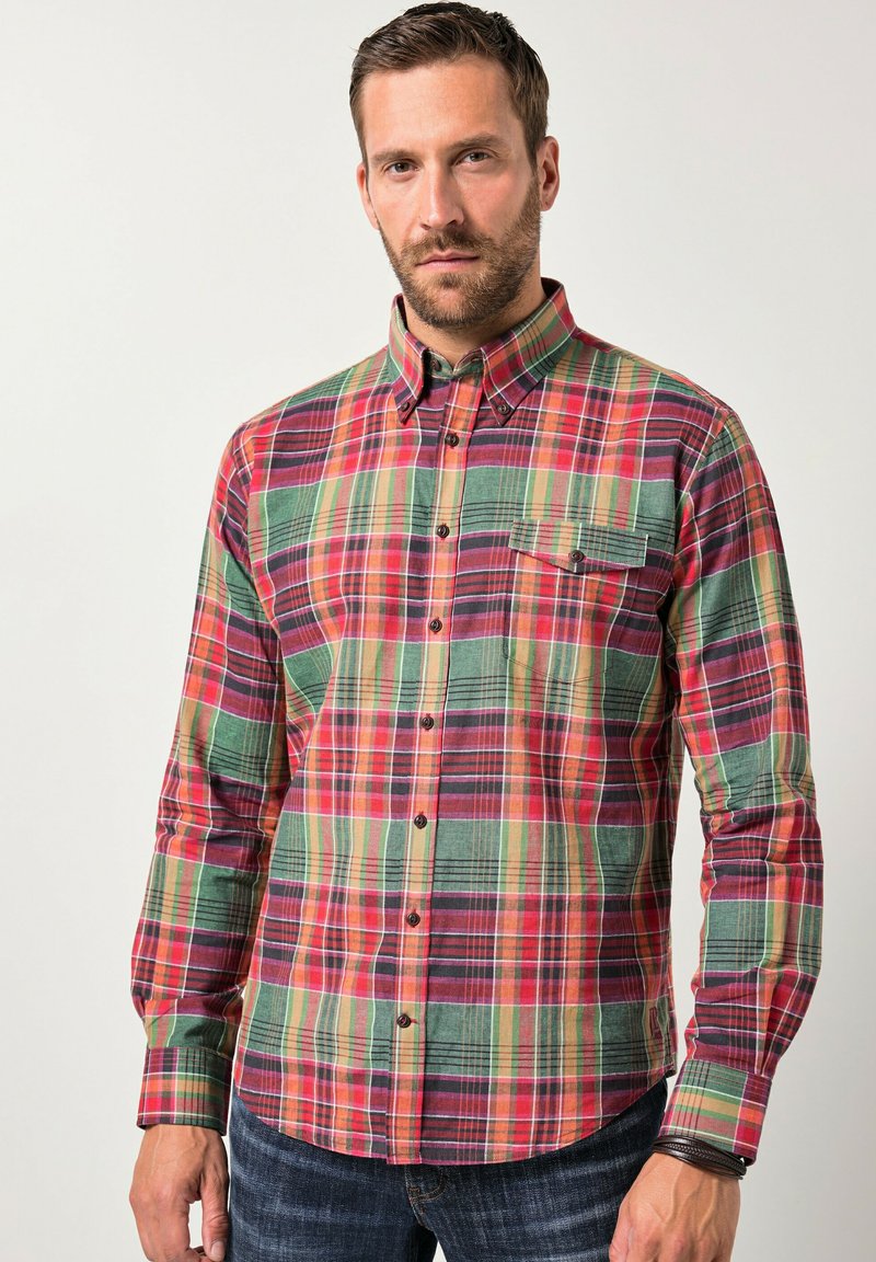 Uomo che indossa una camicia a quadri multicolore rossa, verde e arancione con bottoni e jeans scuri, in piedi davanti a uno sfondo chiaro e uniforme.