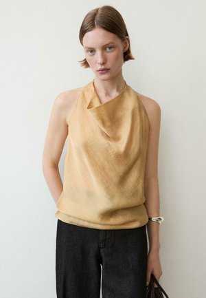 Ung kvinde med kort hår iført en ærmeløs beige draperet top og sorte bukser, står mod en lys, ensfarvet væg.