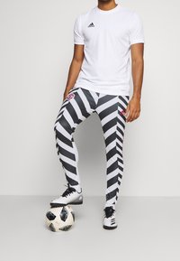 Man i vit Adidas-tröja och svart-vita randiga byxor som står med en fot på en svart och vit fotboll.