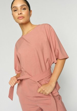 Žena oblečená v voľnom ružovom topu a nohaviciach si na pás viaže široký opasok, na neutrálnom svetlom pozadí.