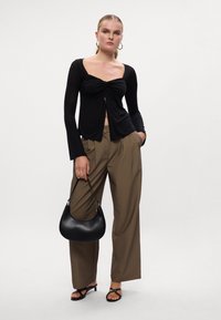 Crna majica dugih rukava s prednjim zavrtnjem, uparen s taupe širokim hlačama i crnim slingback sandalama, držeći zakrivljenu crnu torbu.