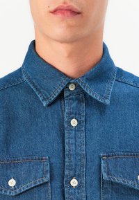 Denim košulja u plavoj boji s oštrim okovratnikom, dva džepa na prsima i bijelim gumbima. Ima vidljivo šivanje i blagu teksturu.
