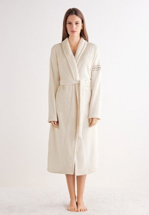 Woman standing barefoot wearing a cream-colored, long bathrobe tied at the waist with long sleeves and a shawl collar.