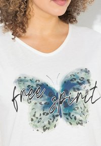 Mujer vistiendo una camiseta blanca de cuello en V con una mariposa azul y verde y el texto "espíritu libre" en el pecho.