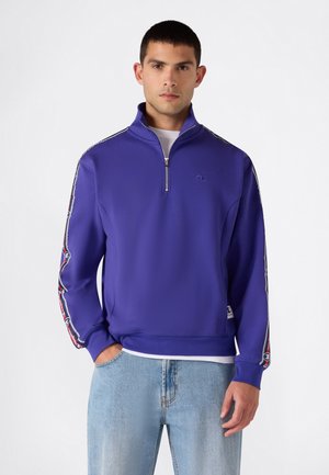 Joven con sudadera morada con cremallera hasta el pecho con rayas de logo en las mangas y vaqueros azul claro, de pie con una mano en el bolsillo.