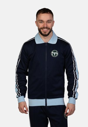 Navy zip-up jacket with light blue collar and cuffs. Features white stripes down sleeves and a circular logo on the chest. Smooth fabric texture.