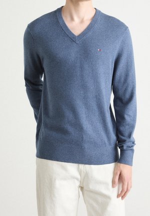 Homme portant un pull bleu col en V avec un petit logo Tommy Hilfiger sur la poitrine et un pantalon beige clair, debout devant un fond uni.