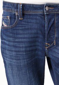 Jeans din denim albastru închis, cu un design clasic cu cinci buzunare, cusături din cupru, închidere cu un buton metalic și o textură ușor decolorată.