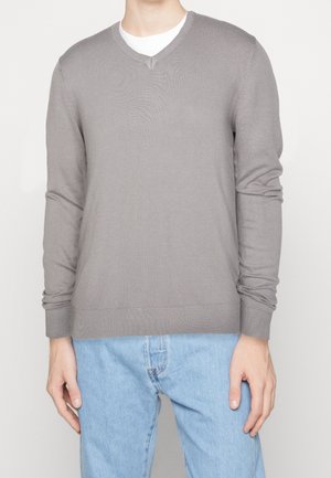 Grå strikket sweater med V-hals, lange ærmer og ribbede manchetter, båret over en hvid T-shirt, parret med lyseblå jeans.