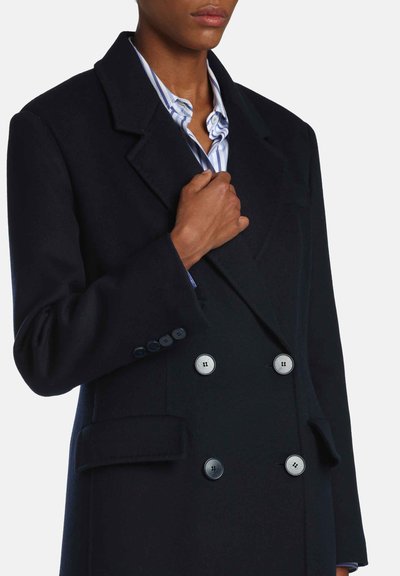 Manteau bleu marine foncé croisé avec de grands boutons gris, coupe ajustée, cols crantés et tissu texturé. Chemise blanche à rayures bleues visible.