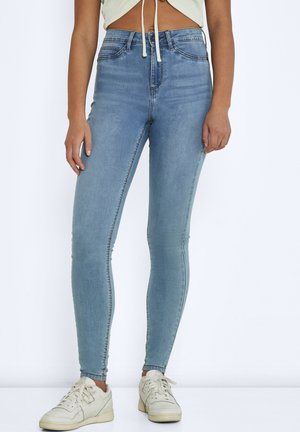 Jeans skinny en denim bleu clair, taille haute avec une silhouette ajustée. Présente un dégradé subtil et des coutures latérales. Porté avec des baskets blanches.