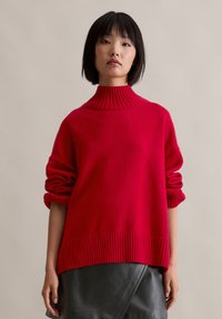 Roter gestrickter Pullover mit hochgeripptem Kragen, weiten Ärmeln und strukturierter Oberfläche, kombiniert mit einem schwarzen Lederrock.