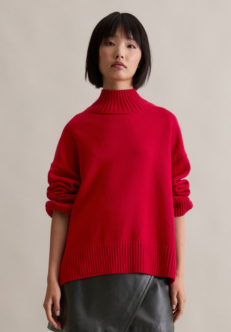 Roter gestrickter Pullover mit hochgeripptem Kragen, weiten Ärmeln und strukturierter Oberfläche, kombiniert mit einem schwarzen Lederrock.