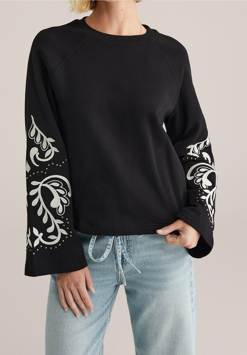 Femme portant un haut noir à manches longues avec broderie florale blanche et détails cloutés sur les manches, associé à un jean en denim bleu clair.