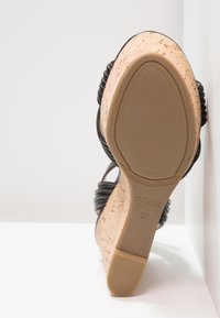 Chaussure à talon en liège noir avec un dessus texturé, dotée de brides tressées et d'une semelle lisse, étiquetée "GUESS" et taille "37" au fond.