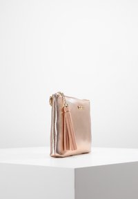 Bolso de mano brillante color oro rosa con un gran tassel a juego colgando de una cadena dorada sobre un pedestal blanco contra un fondo liso.