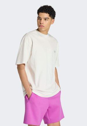 Ung mand med krøllet hår iført en løs hvid t-shirt og lyserøde shorts med hænderne i lommerne, der kigger til siden.