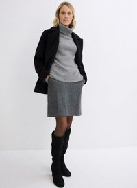 Maglione grigio a collo alto con dettagli sottili, indossato sotto un cappotto nero, abbinato a una gonna grigia e stivali neri sopra il ginocchio.