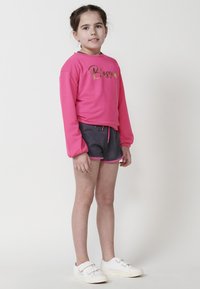 Sweatshirt à manches longues rose avec le mot « Blossom » en lettres dorées, associé à un short gris avec des accents roses. Porté avec des baskets blanches.