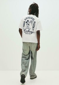 Tricou alb cu un grafic reprezentând pe Mickey Mouse ținând o felie de pizza, cu textul: "Pizza Mickey's, adevărat original, din 1928." Blugi bleu deschis.