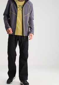 Homme portant une veste zippée grise par-dessus un t-shirt vert à motif, un pantalon noir et des chaussures de plein air noires, debout devant un fond uni.