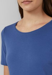 Kobieta w jednolitym, niebieskim, krótkim rękawie koszulce z okrągłym dekoltem, skróconej, aby pokazać szyję, ramiona i górną część klatki piersiowej.