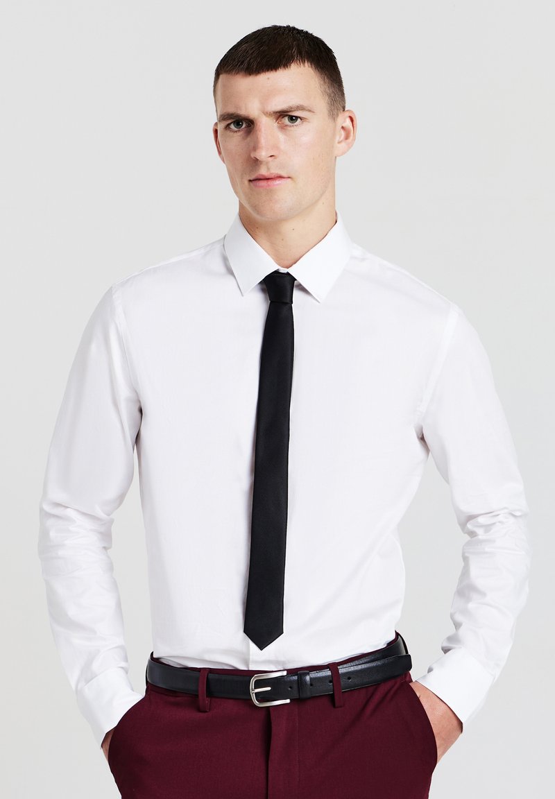 Uomo con capelli corti castani, indossa una camicia bianca, una cravatta nera, una cintura nera e dei pantaloni rosso scuro, in piedi con le mani nelle tasche.