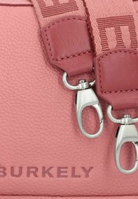 Borsa a tracolla in pelle rosa con texture, dotata di hardware argentato e una tracolla intrecciata con un motivo ripetuto. Il nome del marchio "BURKELY" è impresso.