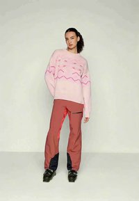 Γυναίκα με σκούρα κατσαρά μαλλιά που φοράει ένα ροζ μοτίβο πουλόβερ, κόκκινα παντελόνια σκι με μαύρες και κόκκινες λεπτομέρειες και μαύρες μπότες σκι, σε απλή φόντο.