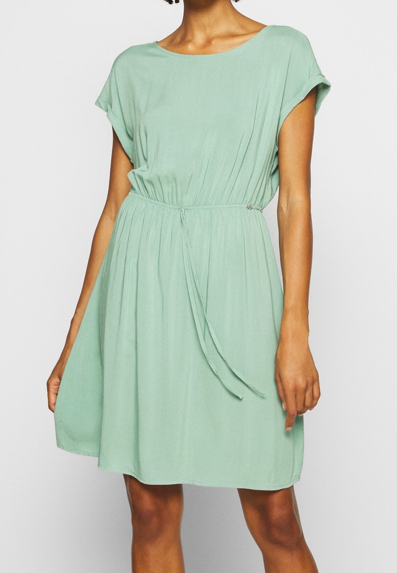 Femme portant une robe vert clair à manches courtes, longueur genou, avec une taille froncée et une ceinture nouée sur un fond uni.
