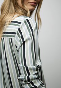 Femme blonde portant une chemise à manches longues avec des rayures verticales en bleu, vert, blanc et noir, vue de profil sur un fond uni.
