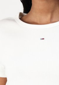 Balta juostuota marškinėliai su apvaliu kaklu ir trumpomis rankovėmis. Krūtinės srityje yra mažas, siuvinėtas logotipas raudonos, mėlynos ir baltos spalvų.